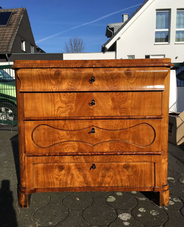 Holzkommode mit vier Schubladen, die auf einem gepflasterten Weg im Freien steht, mit Häusern im Hintergrund.