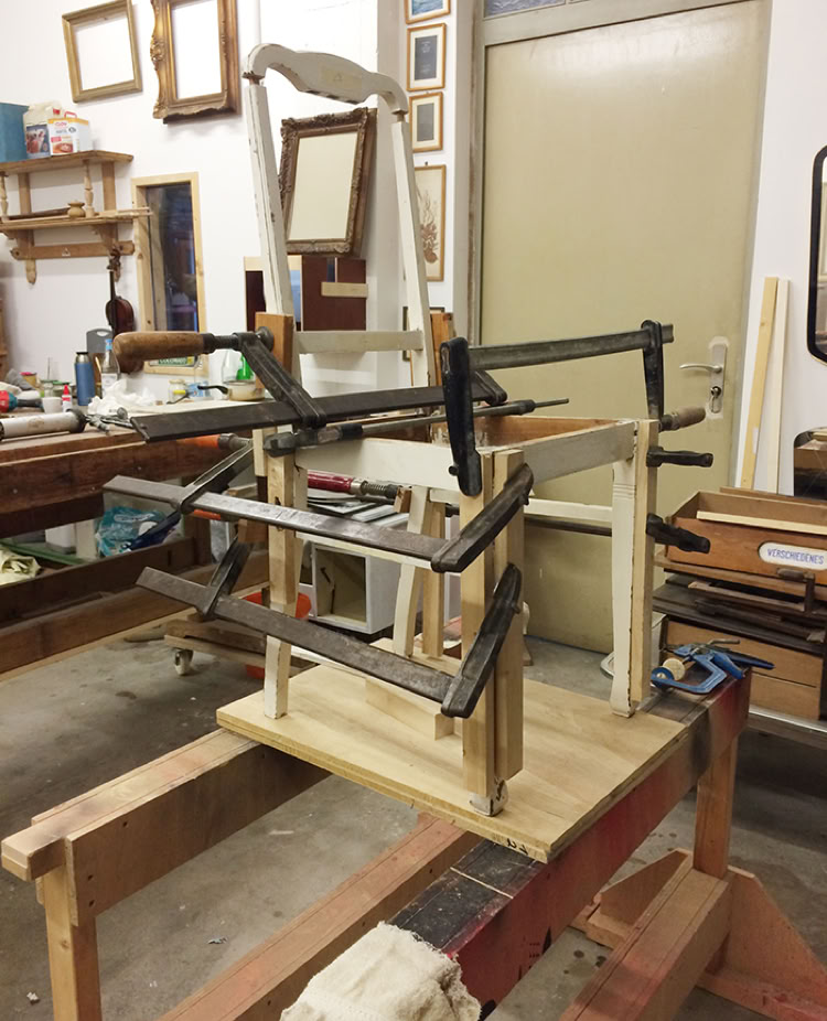 Ein Holzstuhl in einer Werkstatt wird mit mehreren Schraubzwingen repariert. Im Hintergrund sind Werkzeuge, Regale und Bilderrahmen zu sehen.