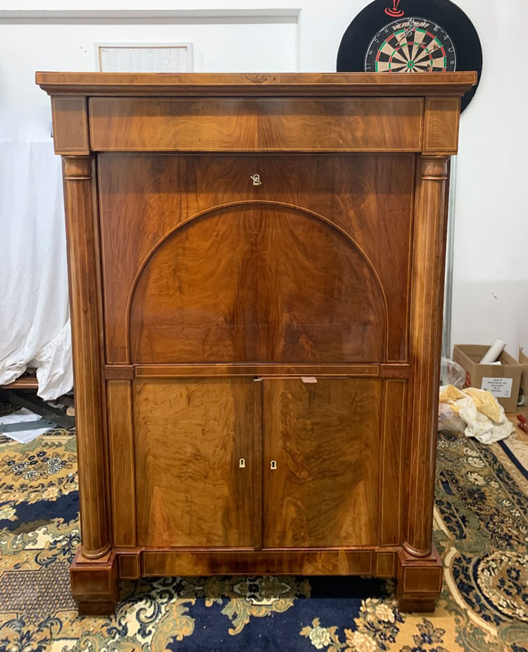 Ein hölzerner Schrank mit zwei Türen und einem gewölbten Design in der Mitte, steht auf einem gemusterten Teppich. Im Hintergrund befindet sich eine Dartscheibe an der Wand.