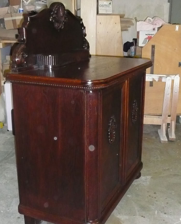 Dunkler Holzschrank mit dekorativem Aufsatz, in einem Raum mit Kartons und Holzmöbeln im Hintergrund.